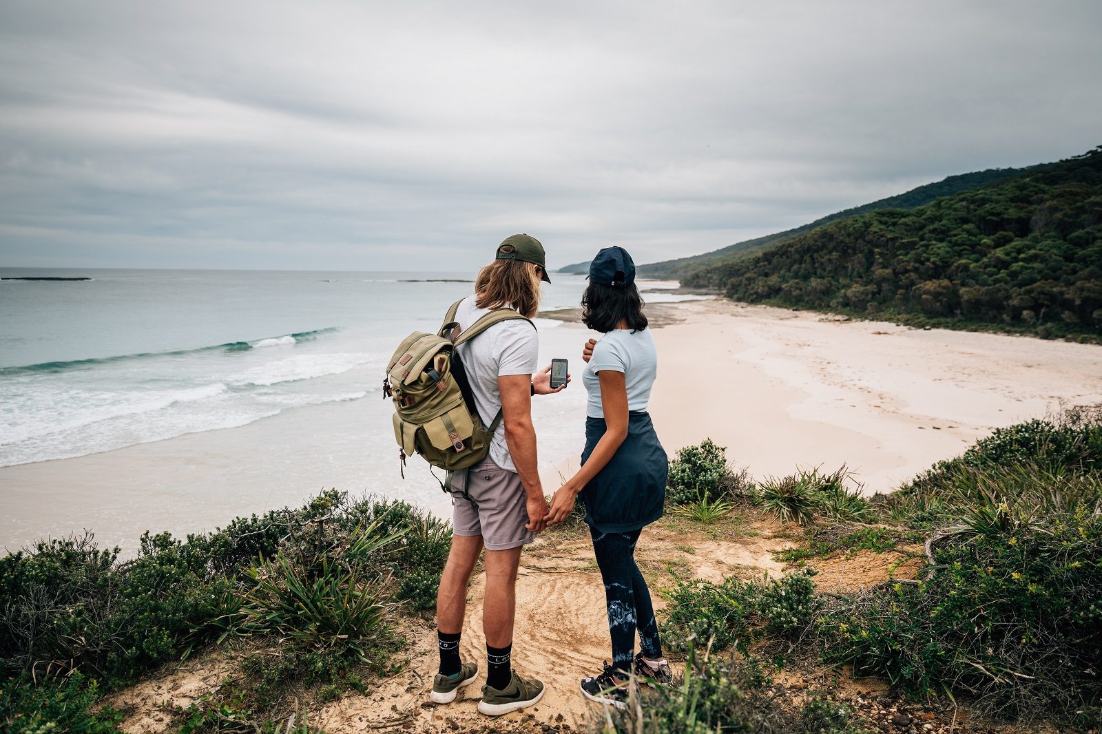 A couple holding hands looking at their mobile device with the view of the beach in front of them at Murramarang National Park. Photo: Melissa Findley/DPIE