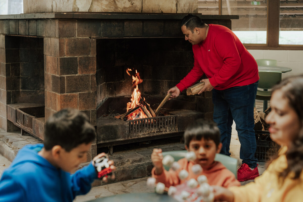 Two children with marshmallow skewers and a man tending to fire in a fireplace. Barmah Dining hall, Wombeyan Karst Conservation Area. Photo credit: Remy Brand / DCCEEW