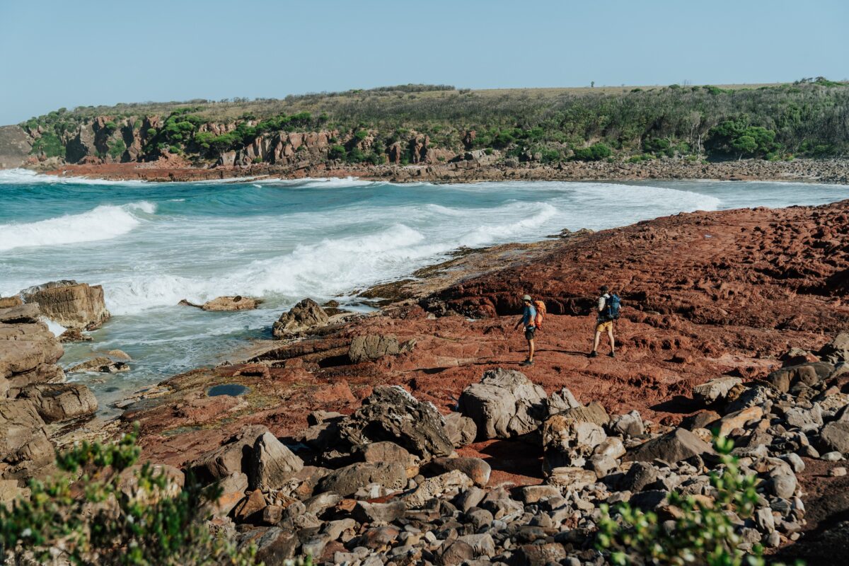Two walkers cross the red rock platform at Hegartys Bay on the Light to Light Walk. Photo credit: Remy Brand/DCCEEW