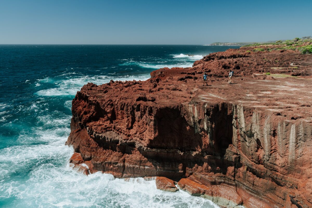 Two walkers on a red rock platform on the Light to Light Walk. Photo credit: Remy Brand/DCCEEW
